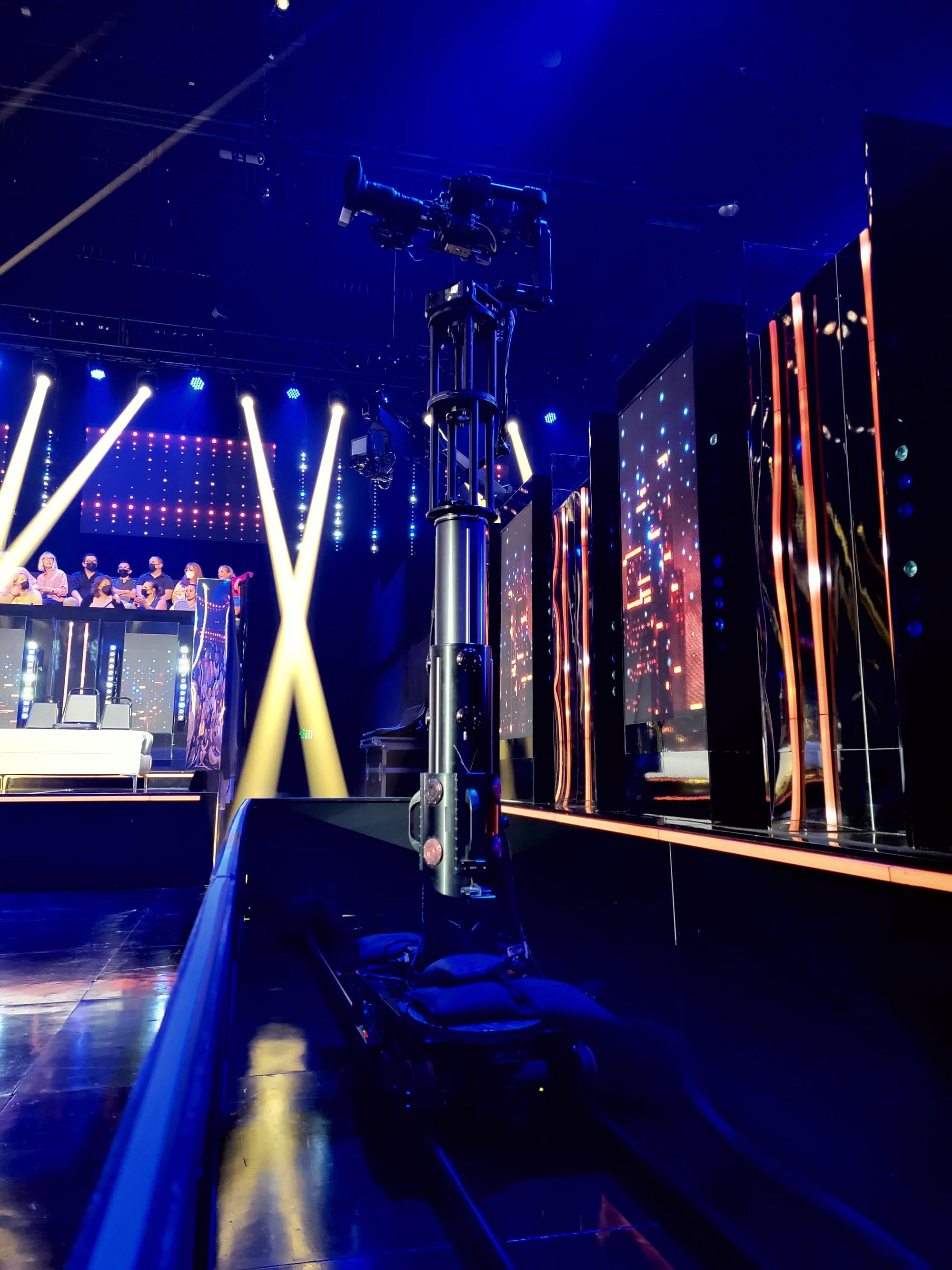Telescoping towercam camera system on a blue-lit broadcast stage with an audience and light beams